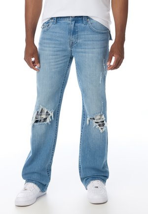 Person trägt hellblaue zerrissene Jeans mit schwarz-weiß karierten Stoffflicken hinter den Knie-Rissen und weiße Sneakers.