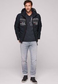 Giacca nera imbottita con zip e cappuccio, caratterizzata da design testuali ricamati. Indossata sopra un maglione in maglia scura e jeans azzurri chiari. Scarpe nere.