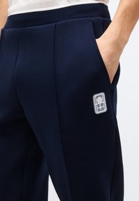 Κοντινό πλάνο ατόμου που φοράει μπλε παντελόνι με το χέρι στην πλαϊνή τσέπη και ένα μικρό ορθογώνιο λογότυπο κοντά στην τσέπη.