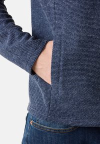 Granatowy sweter o teksturowanej powierzchni, z poziomymi prążkami; posiada przednią kieszeń i dopasowane mankiety. Wykonany z miękkiego, elastycznego materiału.