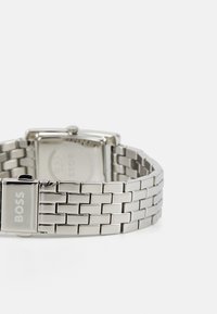 Reloj de pulsera de acero inoxidable plateado con caja rectangular, correa de malla y logo de la marca grabado en el cierre.