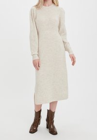 Beige stickad midikjol med lång ärm och rund halsringning, med ribbad textur och en sidofålla. Stylandes med bruna klackstövlar.