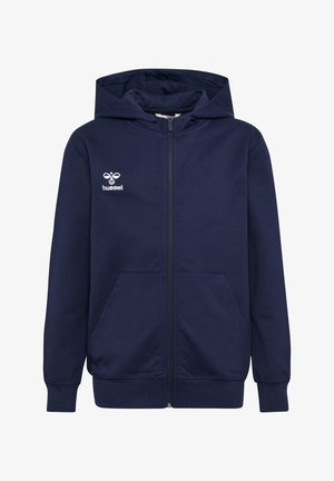 To børn står, iført mørkeblå hættetrøjer; den ene i hvid skjorte og grå bukser med hvide sneakers, den anden i sort skjorte og bukser med sorte sko.