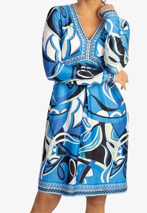 Vrouw die een blauw-wit-zwart jurkje met lange mouwen draagt met abstracte geometrische patronen en een V-hals met decoratieve bies.