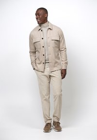 Beige overhemdjack met knopsluiting en twee borstzakken, gecombineerd met een lichtbeige coltrui en bijbehorende broek. Bruine sneakers maken de look compleet.
