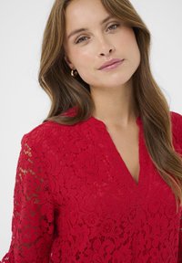 Bluză roșie din dantelă cu un design în formă de V, cu motive florale și mâneci transparente. Prezintă un material texturat și detalii fine din dantelă.