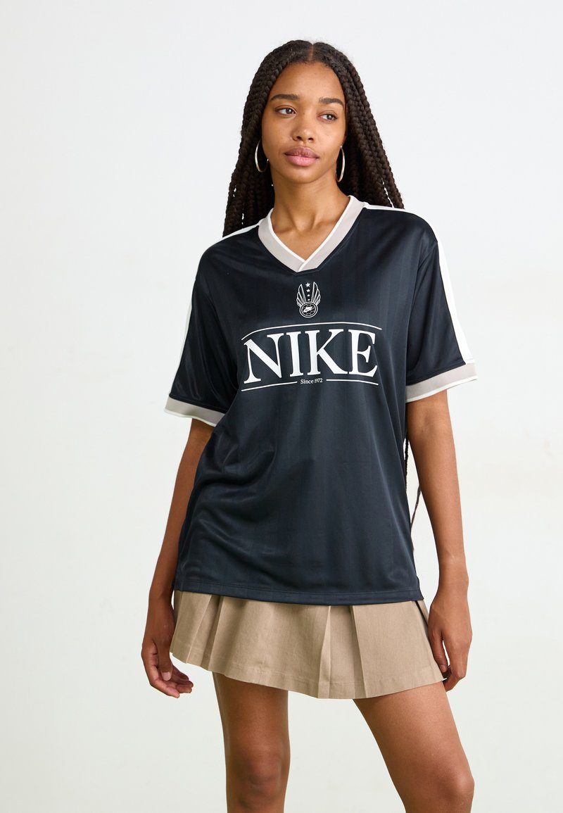 Svart Nike-bluse med hvite detaljer, kort erm, og v-halsdesign. Har en fremtredende logo på forsiden. Teksturen ser jevn ut.