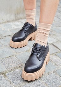 Chaussures noires en cuir avec une semelle épaisse en cuir tan. Elles présentent un bout arrondi, un design à lacets et des chaussettes à motif tan texturé au niveau de la cheville.