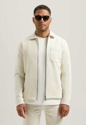 Helle cremefarbene Zip-Jacke mit Kragen, Fronttasche und langen Ärmeln, getragen über einem weißen T-Shirt und passenden Hosen.