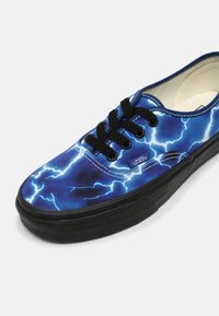 Niebieskie trampki z płóciennego materiału z wzorem błyskawicy, czarnymi sznurowadłami i czarną gumową podeszwą. Widoczny detal logo z boku.