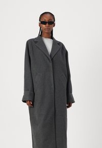 Dlouhý šedý vlněný kabát s klopovým límcem, oversized střihem, širokými manžetami a bočními kapsami. Nosený přes šedý top a doplněný černými slunečními brýlemi.