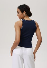Marineblå ribbet tanktop med hvit kontrastsøm og en tettsittende design. Brukt sammen med hvite høyhåndte bukser med baklommer.