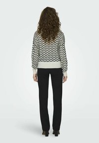 Maglione lavorato a maglia in bianco e nero con motivo a zigzag, maniche lunghe e orlo a coste. Abbinato a pantaloni neri, modello girato di spalle.
