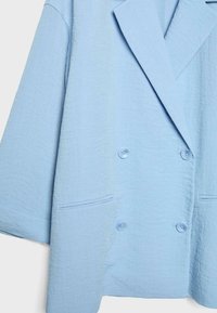 Blazer croisé bleu clair avec larges revers, deux poches avant, et boutons assortis bleus sur tissu texturé.