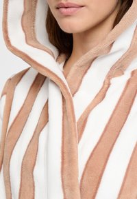 Kuklový plášť vyrobený z měkkého, plyšového materiálu s střídajícími se krémovými a pískovými pruhy, s volným střihem a viditelnými švy.