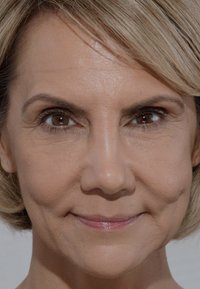 Gros plan sur le visage d'une personne avec un teint clair, une texture lisse et des rides subtiles. Les cheveux sont courts et blonds, encadrant le visage.