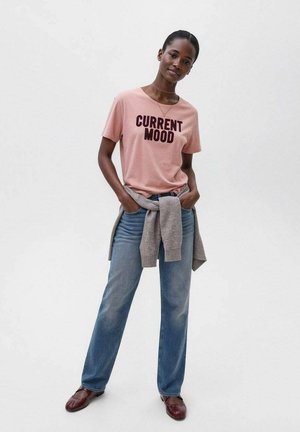 T-shirt rose à manches courtes avec le texte noir en gras « ÉTAT D'ESPRIT ACTUEL », associé à un jean droit bleu clair et un pull gris noué à la taille.