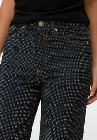 Tamne traperice tamne boje s uzorkom malih metalnih zakovica po cijeloj površini, s prednjim kopčanjem na dugme i toniranim detaljima šivanja.