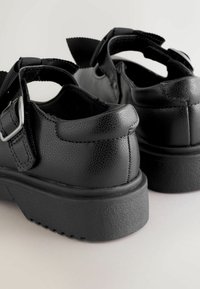 Zwarte schoenen van gestructureerd synthetisch materiaal, met een dikke rubberen zool, verstelbare riem en een afgeronde achterkant met een trekriempje.