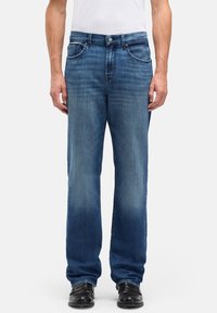 Jeansy z denimu w średnim niebieskim kolorze o prostym kroju nogawek, wyposażone w przednie kieszenie, zapięcie na guziki i subtelne przetarcia. Noszone z czarnym obuwiem.