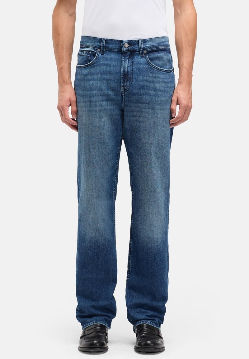 Jeansy z denimu w średnim niebieskim kolorze o prostym kroju nogawek, wyposażone w przednie kieszenie, zapięcie na guziki i subtelne przetarcia. Noszone z czarnym obuwiem.