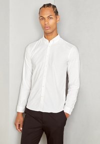Camisa branca de mangas longas com colarinho, apresentando uma textura suave e um design ajustado. Usada com calças pretas.
