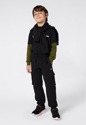 Pantaloni sportivi neri con tasche cargo, stratificati con una maglietta a maniche lunghe olivette sotto una maglietta a maniche corte nera, e sneakers nere con dettagli rossi.