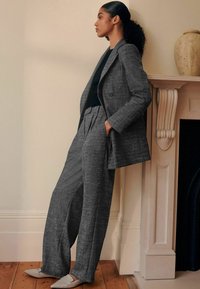 Donna con capelli ricci, in un completo a quadri grigio e top nero, in piedi di lato con le mani nelle tasche vicino a un caminetto bianco.