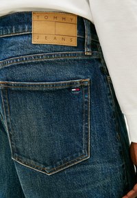 Pantalones vaqueros de mezclilla con un lavado azul oscuro, que presentan una parche de cuero marrón etiquetado como "TOMMY JEANS" y acentos en rojo, blanco y azul cerca del bolsillo.