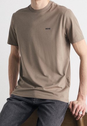 Homme portant un t-shirt marron à manches courtes avec le logo "BOSS", jean foncé et bague en argent, assis sur une chaise en bois avec une main reposant dessus.