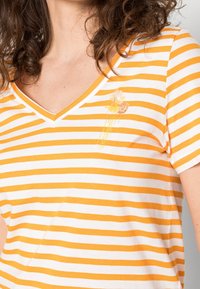 Žena v bílém a oranžovém pruhovaném V-neck tričku s malým květinovým vzorem na hrudi, viditelné kudrnaté hnědé vlasy.