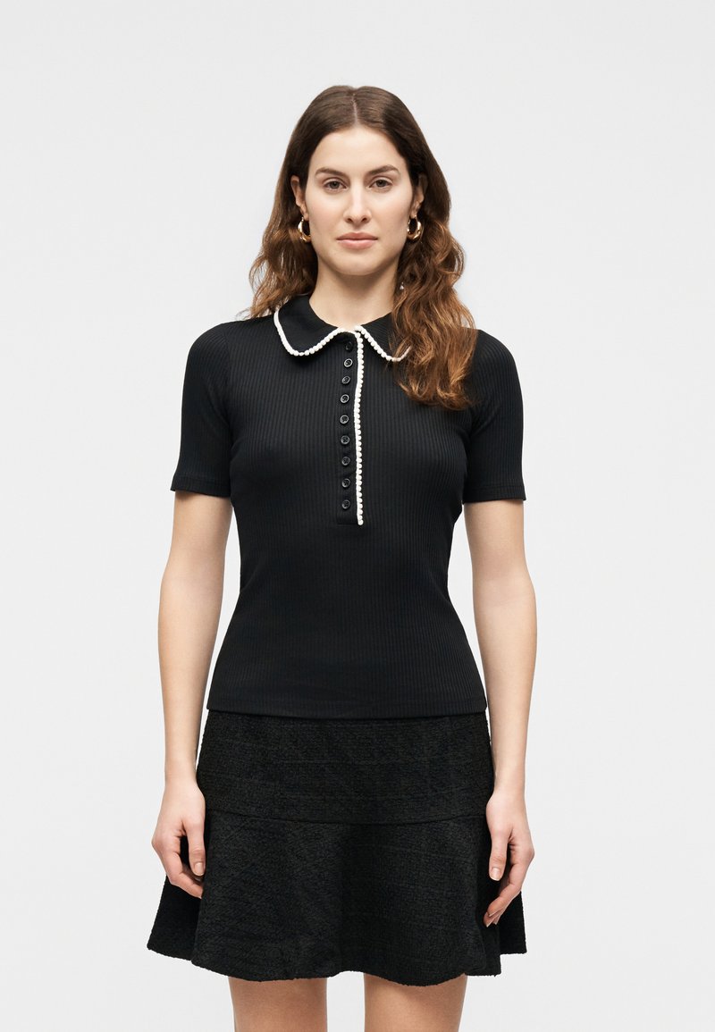 Femme aux longs cheveux bruns portant un haut noir à manches courtes avec un col et des boutons bordés de blanc, assorti à une jupe noire texturée.