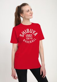 Young woman with brown ponytail wearing a red "Shibuya 1961 Tokyo Baseball" t-shirt and black pants, looking upward to right.
