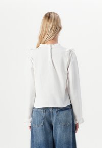 Donna con lunghi capelli biondi che indossa una blusa bianca a maniche lunghe con dettagli in pizzo e jeans di denim blu, vista di spalle su uno sfondo neutro.