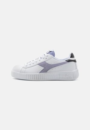 Zapatilla blanca con acento de ante púrpura y talón negro. La suela de goma texturizada presenta estrías verticales. Cierre con cordones y lengüeta acolchada.