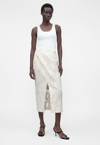 Γυναίκα με λευκό τοπ, κρεμ φλοράλ midi φούστα με άνοιγμα μπροστά και μαύρα πέδιλα με τακούνι στέκεται μπροστά σε απλό φόντο.