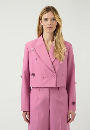 Kvinde med langt lyst hår iført en cropped lyserød dobbeltradet blazer dekoreret med små blomsterapplikationer og matchende bukser, står stille.