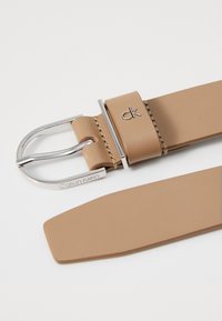 Cintura in pelle tan di Calvin Klein con fibbia in metallo argentato, dotata di passante con logo in rilievo e texture liscia. Punta piatta per un facile fissaggio.