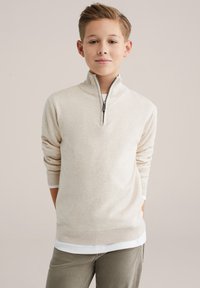 Šviesiai gelsvas megztinis su pusiau užtrauktuku apykaklėje, su ribinėmis rankogaliais ir apačia. Dėvimas virš baltos marškinių, turi lygią tekstūrą ir aptemptą siluetą.