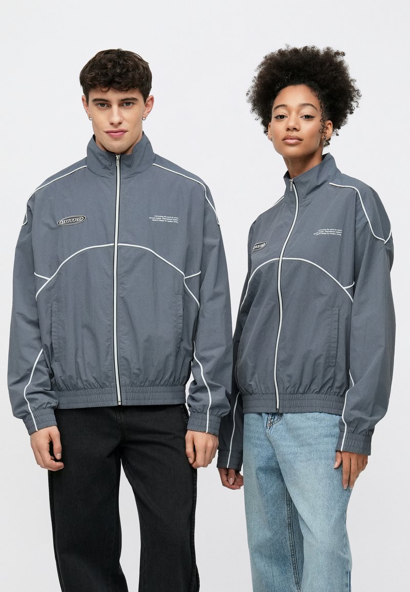 Grå vindjakker med hvite pipedetaljer, glidelås foran og elastiske mansjetter. To modeller har dem på, stylet med kontrasterende svarte og lyseblå jeans.