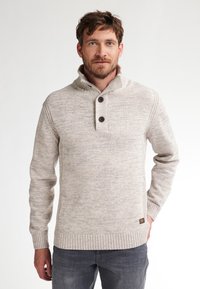 Uomo con barba che indossa un maglione lavorato a maglia beige con due bottoni e jeans grigio scuro su uno sfondo bianco semplice.