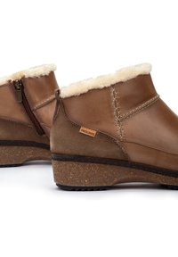 Botas de tobillo de cuero marrón con un suave forro de piel de piel, detalles cosidos, cremallera lateral y una suela de cuña de corcho texturizada. Etiqueta de la marca visible.