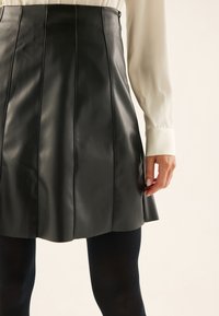 Gonna nera in ecopelle con pieghe, caratterizzata da una vita alta e cuciture a contrasto, abbinata a una blouse bianca a maniche lunghe e collant neri.