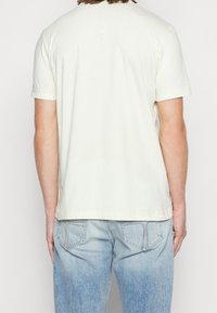 Kremowa bawełniana koszulka z krótkim rękawem, okrągłym dekoltem i obszytym dołem, noszona z jasnoniebieskimi, wyblakłymi dżinsami.