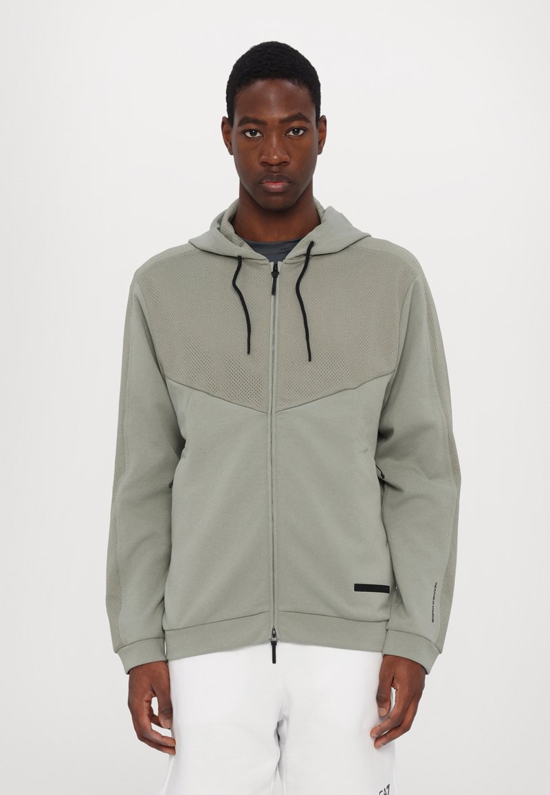 Sudadera gris con cremallera y textura de malla en la parte frontal superior. Cuenta con una capucha ajustable y bolsillos con cremallera a los lados.