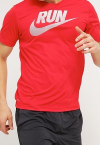 Czerwona koszulka sportowa z napisem "RUN" w białym kolorze oraz logo Nike. Krótkie rękawy, dopasowany krój, gładka tekstura. Połączona z ciemnymi szortami.