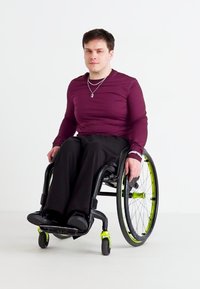 Ένα άτομο σε έναν μαύρο αναπηρικό καρότσι με πράσινες λεπτομέρειες, φορώντας μια μαροουν μπλούζα με μακριά μανίκια και μαύρο παντελόνι, καθισμένο μπροστά σε λευκό φόντο.