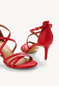 Sandales à talons hauts rouges en cuir lisse, dotées de multiples brides entrecroisées et d'une sangle de cheville à boucle. Talon fin avec un dos fermé.