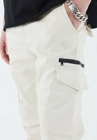 Beige cargo bukser med en tekstureret finish, der har en lynlås-lomme, en side cargo-lomme og broderet logo-detalje.