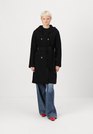 Manteau noir à double boutonnage avec capuche, doté d'une ceinture à nouer, de grands boutons et de deux poches avant, associé à un jean évasé et des chaussures rouges.
